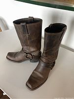 Right and left Frye brown leather harness boots shown upright on white surface, visible leather wear and creases.