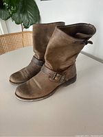 Pair of full brown leather boots seen at an angle showing outer side and back, placed on floor with plant background