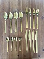 16 pieces of gold colour cutlery including spoons, forks, knives and teaspoons laid out on a wooden surface.