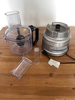 Complete food processor set on wooden table including motor base, clear plastic bowl with handle and lid, two plastic food pushing tubes, and metal chopping blade.