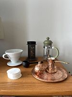 Photo showing all coffee equipment: white ceramic V60 dripper, black AeroPress, green-handled French press, black scoop, stack of paper filters, copper ibrik with lid on a round copper tray.