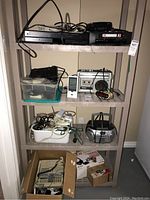 Shelf containing multiple electronics including a JVC DVD player, VHS players, a radio cassette player, containers of cables, and remote controls.