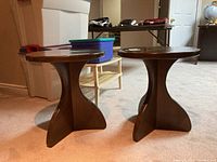 Pair of matching wooden side tables shown from the side at an angle, revealing round tops and curved bases.