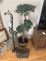 Photo showing full view of carved wood giraffe next to faux plant in planter and metal basket.