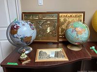 Two world globes, two gold decorative wall maps, framed black and white vintage print, and a small music box figurine on wooden table