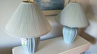 Two matching light blue ceramic lamps with pleated fabric shades placed side by side on a white surface.
