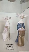 Two Lladro porcelain figurines, one kneeling in blue dress with prayer stand, one seated in white dress holding object.