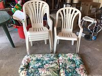 Five white resin stacking outdoor chairs with armrests and slatted backs stacked together, positioned near a floral-patterned padded chair cushion with green, pink, and white colors; photo taken to show entire set.
