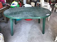 Full view of the green plastic resin patio table with oval top and four legs, showing the overall shape and condition with visible scratches and dirt on the surface.