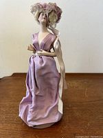 Full view of the 10-inch porcelain half-doll figure dressed in a purple tunic with lace on the hat and jointed arms.