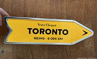 Arrow-shaped yellow tin sign with black text, showing brand Veuve Clicquot, TORONTO, and distance to Reims 6,095 km. Photo shows the sign flat on a wooden floor.