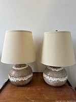 Pair of Maurice Chalvignac pottery lamps on wooden surface with beige fabric shades unlit.