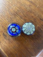 Two vintage mini Murano millefiori art glass paperweights on wooden surface, showing intricate millefiori floral patterns, one bright blue and one cream-green.