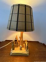 Full table lamp with carved pine wood base depicting Quebec habitant scene and beige brown grid lampshade, lit to show working condition.