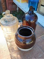 Three items on wooden surface outdoors: clear embossed glass jug, brown glass jug, brown ceramic pot.
