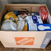 Cardboard Home Depot Petite box filled with multiple light bulbs in original packaging including yellow, blue, and red boxes.