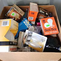 Top-down view of a large cardboard box filled with various boxed light bulbs including GE and Sylvania brands, different wattages and types visible.