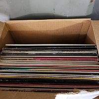 Cardboard box filled with a stacked assortment of LP vinyl records showing the top edges and spines of album covers.