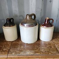 Front view of three vintage stoneware moonshine jugs with glazed brown tops and white bodies on wooden surface.