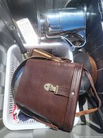 Photo showing a brown leather binocular case with a strap lying on top of a white basket containing various items including a brass pan and other metal objects.