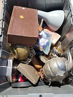 Storage bin containing various kitchenware items including a metal teapot, white funnel, glassware, electrical lamp with cord, and miscellaneous small items.