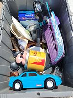 View inside plastic bin showing assortment of items including toys, action figures, a ceramic spoon, a Verbatim CD-R pack, a model racing car, a blue toy car, a Mickey Mouse figure, and a yellow tin.