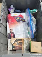 View inside storage bin showing various items including papers, VHS tape in a bag, boxed item, and an action figure partially visible