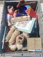 Top-down shot of a storage bin filled with various items including a large doll, stuffed animals, books, and glassware.