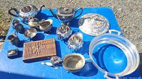 Wide photo showing multiple copper, brass, and silver plated vintage items including tea sets, trays, bowls, and decorative boxes on a blue surface outdoors.