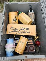 Close top-down view of storage bin containing wooden containers with Asian inscription, ceramic vase, brass items, and wooden box with Asian calligraphy.
