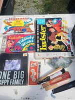 Photo of the storage bin contents showing multiple vintage board games including The Amazing Spider-Man Game with The Fantastic Four, the Twister game, Combination Tiddledy Winks, along with wooden hand fans, bags of marbles, and miscellaneous papers.