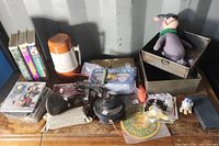 Photo showing assorted items including VHS tapes, DVDs, stuffed toy, toy gun, coffee thermos, and small items on a table.