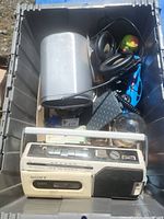 Plastic storage bin containing Sony cassette player, metal tin, headphones, and assorted small items