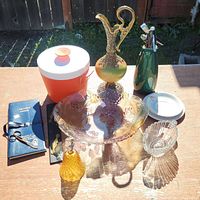 Overview of collectible lot items on table outdoors including glassware, container, pitcher, soda siphon, bell, dishes, and booklet.