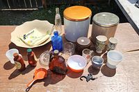 Photo of assorted glassware, bottles, tins, cups, and small kitchenware items arranged on a wooden surface outdoors.