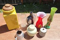 Wide shot showing all collectible vases, jars, and the yellow ceramic container on a wooden surface outdoors.