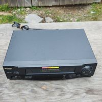 Black Sharp VCR unit showing front and top with visible power cord and cartridge slot.