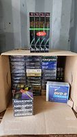 Large cardboard box with multiple sealed VHS tapes stacked inside, a 5-pack of Fujifilm HQ 120 VHS tapes standing upright on top, two small packs of Memorex DVDs at front right, and a single pack of FujiFilm DVD-R disks.