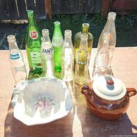 Photo of vintage glass bottles, ceramic pot, and decorative bowl arranged on wooden surface outdoors in sunlight.