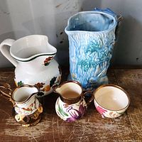 Photo of five Wade pottery creamers and pitchers on wooden table against gray wall, showing overall size and details of hand-painted floral designs and textured surfaces.