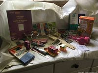 Full lot spread out on white cloth backdrop showing majority of items including pastel paper, crafting tools, colored pencils, plastic butterfly shapes, pink headphones, and kitchen egg cooker box.