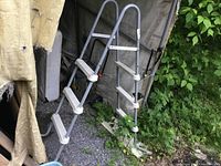 Photo showing 4 foot above ground pool ladder, featuring grey metal frame and white plastic steps with safety handrails, positioned outdoors near wall and greenery.