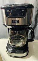 Front view of TRU coffee maker showing control panel, brand logo, and glass carafe.