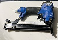 Side view of a blue pneumatic brad nailer with black grip and a label on the magazine strip.