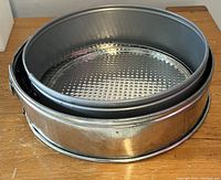Set of three round metal cake tins stacked, top two are standard round tins with textured bases, bottom one has a latch clasp indicating a springform style.