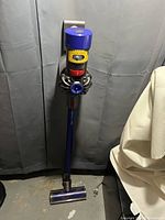 Front view of purple and yellow Dyson stick vacuum mounted on grey wall mount.