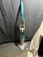 Full view of the teal and white triangular base SteamTek steam mop with upright handle and cord wrapped around the handle.