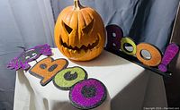Photo of Halloween decorations showing ceramic jack-o-lantern and glitter 'BOO!' sign with purple spider.