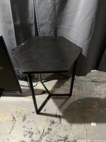 Photo of a black hexagon-shaped plant stand with a metal frame, sitting on a concrete floor with a dark curtain background.