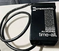 Black rectangular timer unit labeled Intermatic All Weather Time-All with attached black power cable coiled beside it on white surface.